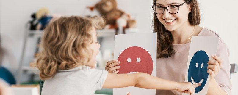 : Docente y alumna en una mesa usando una tarjeta de emociones en una dinámica de apoyo al desarrollo infantil